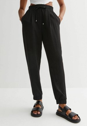 Persona vistiendo pantalones jogger negros con cordón y sandalias negras de punta abierta, de pie con las manos en los bolsillos contra un fondo liso.