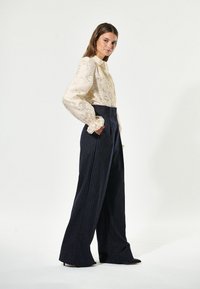 Blusa color crema con motivi floreali testurizzati e polsini a volant, abbinata a pantaloni larghi a righe blu.