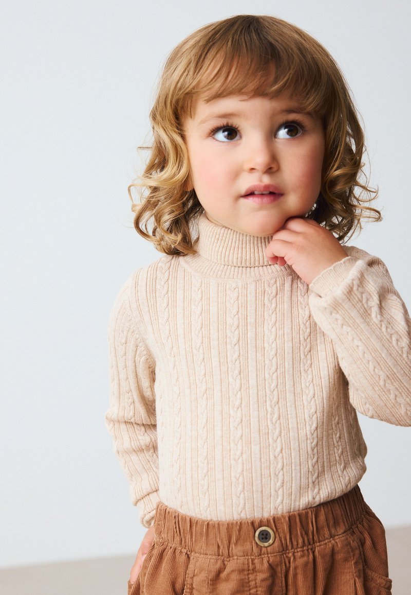 Beige kabelstickad tröja med hög krage, kombinerad med bruna texturerade byxor med elastisk midja och knapprad detalj.