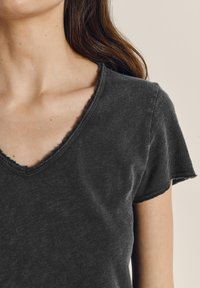 Ženska, oblečena v temno sivo majico kratkih rokavov z izrezom v obliki črke V z neobdelanim robom, ki razkriva ključnico in svetlo kožo s rjavimi lasmi.