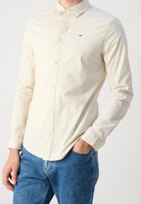 Camicia beige chiaro a maniche lunghe con colletto a punta, chiusura con bottoni e piccolo logo. Indossata con jeans in denim blu. Tessuto dalla consistenza morbida.