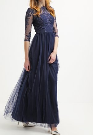 Vrouw in een marineblauwe avondjurk tot op de vloer met doorschijnende mouwen geborduurd met motieven, draagt zilveren hoge hakken en staat voor een witte achtergrond.