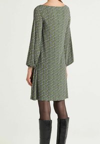 Robe verte avec un motif géométrique en violet et blanc. Manches longues en forme de cloche ; coupe ample ; longueur au genou ; portée avec des bottes noires montant jusqu'au genou.