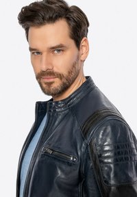 Uomo con barba e capelli stilizzati, che indossa una giacca di pelle nera sopra una camicia azzurro chiaro, guardando con sicurezza verso la telecamera.