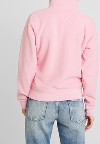 Polarowy welur w jasnoróżowym kolorze z wysokim kołnierzem, ściągaczami przy rękawach i pasie; noszony z jasnoniebieskimi dżinsami, widok z tyłu.