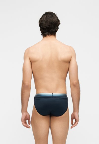 Άνδρας που στέκεται με την πλάτη, φοράει σκούρα μπλε μαγιό με ανοιχτό μπλε ζώνη σε απλό λευκό φόντο.