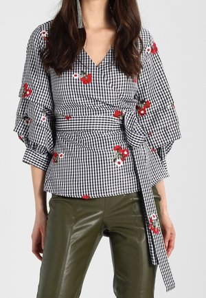 Femme portant un chemisier cache-cœur à carreaux noir et blanc avec broderie florale rouge et pantalon en cuir vert olive.