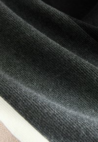 Tmavě šedá látka s texturovaným vzorem, která má podél okraje světle béžový pruh. Materiál vypadá měkce a útulně.