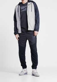 Conjunto deportivo de Nike que incluye una chaqueta gris y negra con cremallera, una camiseta negra, unos pantalones de chándal negros y unas zapatillas gris con acentos coloridos.