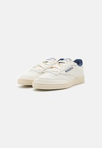 Hvite lær sneakers med perforerte detaljer, beige såle og blå aksenter på kragen. Har flate lisser og Reebok-logo på siden.