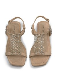 Sandali in suede beige con suola piatta, decorati con un motivo circolare di cristalli lungo la cinghia centrale e una fibbia regolabile alla caviglia.