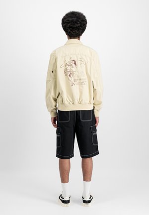 Beige Bomberjacke mit elastischen Bündchen und Saum, die eine bunte Illustration auf dem Rücken zeigt. Kombiniert mit schwarzen Cargo-Shorts und Sneakers.