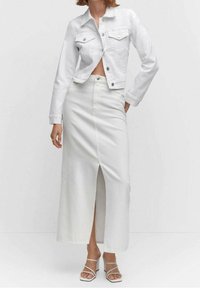 Giacca di denim bianca con due tasche sul petto, abbinata a una gonna bianca al ginocchio con uno spacco frontale, completata da sandali bianchi con cinturini.