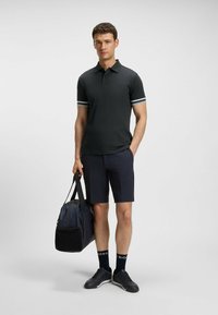 Svart polotröja med vita randiga detaljer, kombinerat med marinblå shorts. Personen håller en mörk duffelväska och har på sig svarta sneakers och strumpor.