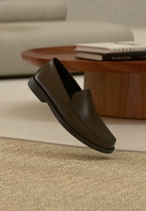 Temno rjavi usnjeni loafer čevelj lebdi nad bež tkanim preprogo z lesenim okroglim kavnim mizico in knjigami v ozadju.