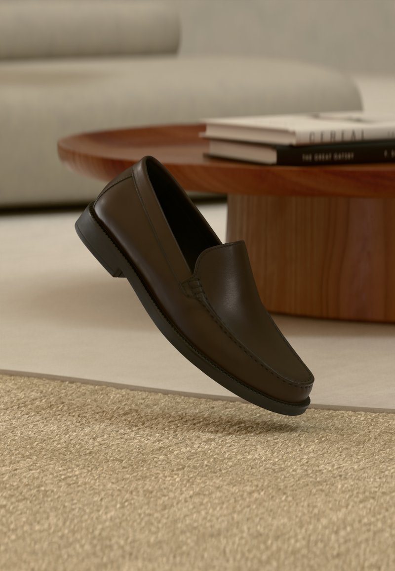 Σκούρο καφέ δερμάτινο μοκασίνι να αιωρείται πάνω από μπεζ πλεκτό χαλί με στρογγυλό ξύλινο τραπεζάκι καφέ και βιβλία στο βάθος.