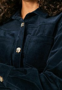 Nainen, jolla on päällä tummansininen corduroytakki suurilla nappilukituksilla ja rintataskuilla, kädet ristissä lähellä vartaloa.