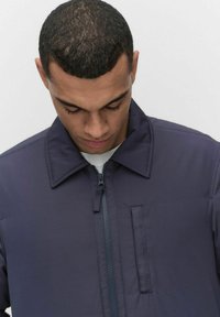 Mörkblå jacka med blixtlås och krage. Har en bröstficka och en slät, något glansig yta.