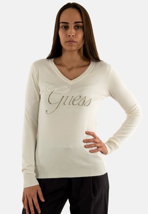Maglione lungo a maniche con scollo a V di colore crema, con logo decorativo "Guess" in strass. Polsini e orlo a costine, texture liscia.