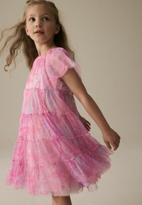 Rosa gestufte Kleid mit Rüschenschultern, versehen mit einem wirbelnden Muster und subtiler Glitzer. Das Material wirkt leicht und luftig.