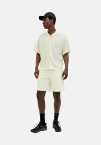 Conjunto de camisa de manga corta amarilla claro con estampado y pantalones cortos, textura de algodón, combinado con gorra negra y zapatos deportivos, en pose de pie.