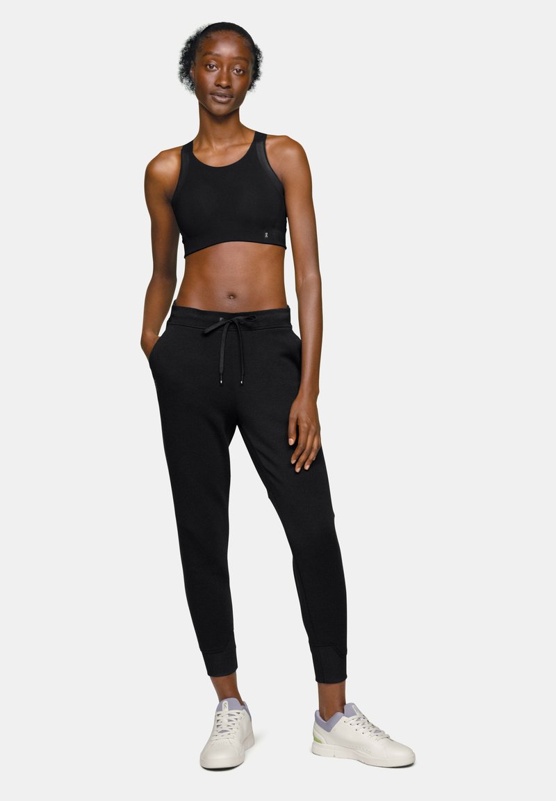 Soutien-gorge de sport noir associé à un pantalon de jogging noir avec une taille élastique et un cordon de serrage. Porté avec des baskets blanches mettant en valeur un accent violet.
