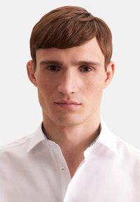 Hvid skjorte med krave i en tekstureret stof, der har en mønstret indre krave. Modellen har kort brunt hår og et neutralt udtryk.