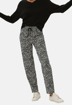 Person trägt schwarzen Pullover, schwarz-weiß gemusterte Hose mit Kordelzugbund und weiße Turnschuhe, steht mit ausgestreckten Armen.