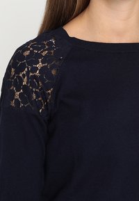 Žena v tmavě modrém svetru s květinovou krajkovou aplikací na rameni a horní části rukávu.