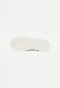 Semelle de chaussure blanche en caoutchouc avec un motif texturé, présentant des rainures en zigzag et un emblème circulaire au centre. Design épuré et minimal.