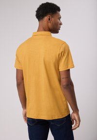 Polo à manches courtes de couleur jaune moutarde, fabriqué dans un tissu doux et texturé. Présente un col classique et un ourlet droit.