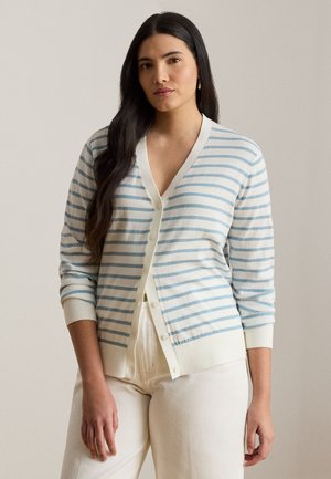 Femme aux longs cheveux foncés portant un cardigan blanc à rayures horizontales bleu clair et un pantalon blanc, debout devant un fond uni.