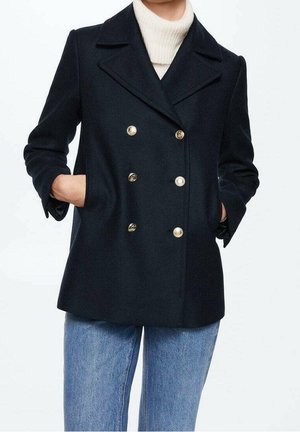 Femme portant un manteau bleu marine à double boutonnage avec des boutons dorés, un col roulé blanc et un jean bleu, les mains dans les poches du manteau.