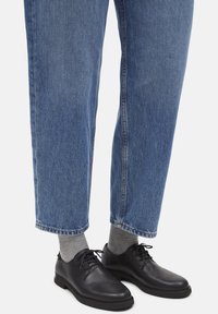 Scarpe di pelle nere con punta rotonda, design con lacci e suole basse, abbinate a calzini grigi chiari e jeans di denim blu con risvolto.