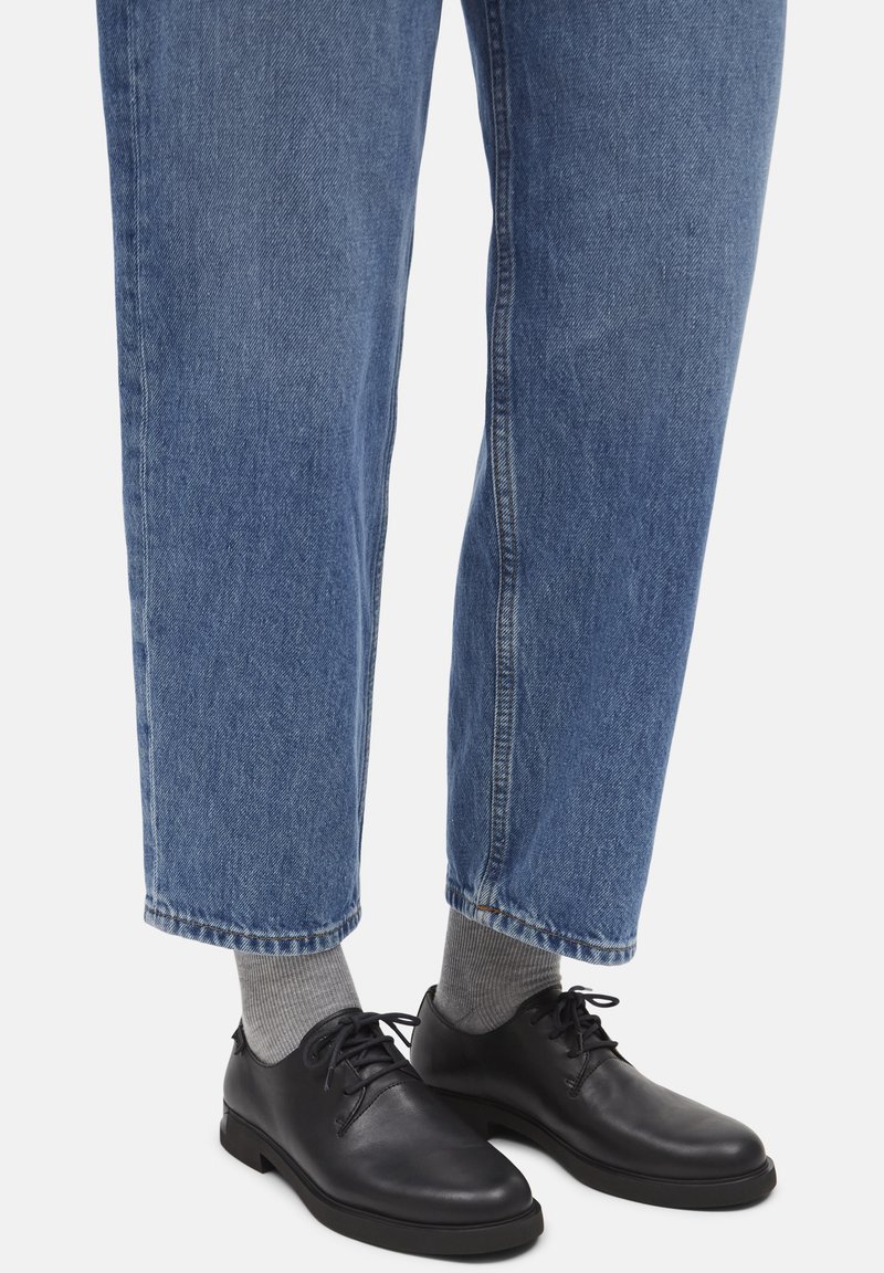 Schwarze Lederschuhe mit runden Zehen, Schnürdesign und flachen Sohlen, kombiniert mit hellgrauen Socken und blauem, umgeschlagenem Denim-Jeans.