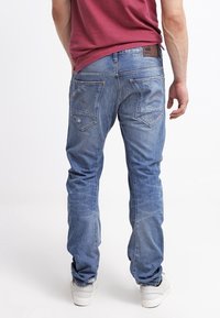 Hombre con jeans azules desgastados y zapatillas blancas, de pie con la espalda hacia el espectador contra un fondo blanco liso.