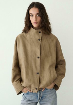 Donna con capelli lunghi e ondulati che indossa una giacca beige a righe sottili, abbottonata e ampia, e jeans azzurri, in piedi con le mani nelle tasche.