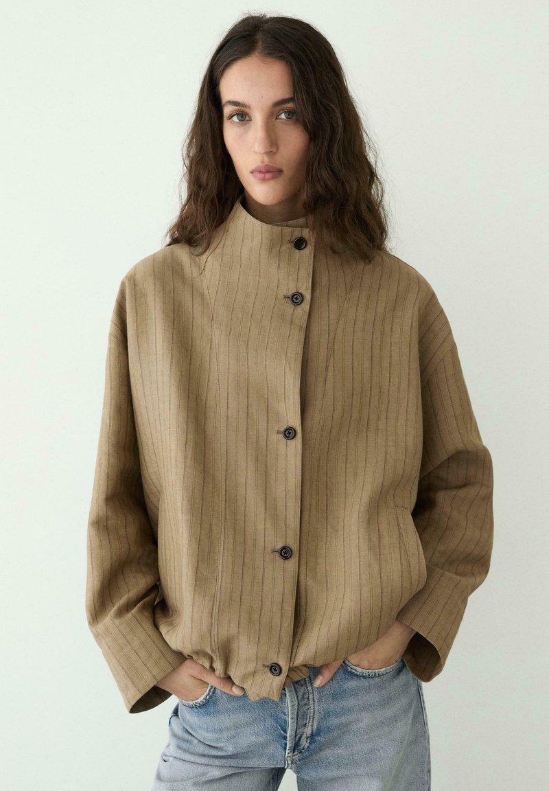 Vrouw met lang golvend haar, gekleed in een losse, beige gestreepte jas met knopen en lichtblauwe spijkerbroek, staand met handen in de zakken.
