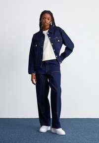 Jaqueta de ganga e calças largas em azul escuro com costuras amarelas; mulher a usar uma t-shirt branca e ténis brancos.