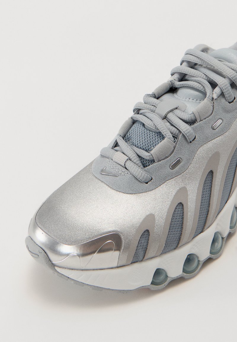 Zapatilla deportiva de color gris claro con una parte superior de plata brillante, detalles de malla, cordones redondeados y una suela translúcida con amortiguación de aire.