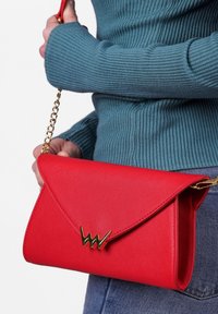 Červená crossbody kabelka s texturovaným vnějším povrchem, trojúhelníkovým klopou a zlatými detaily. Viditelný řetízkový popruh a volný modrý žebrovaný svetr.