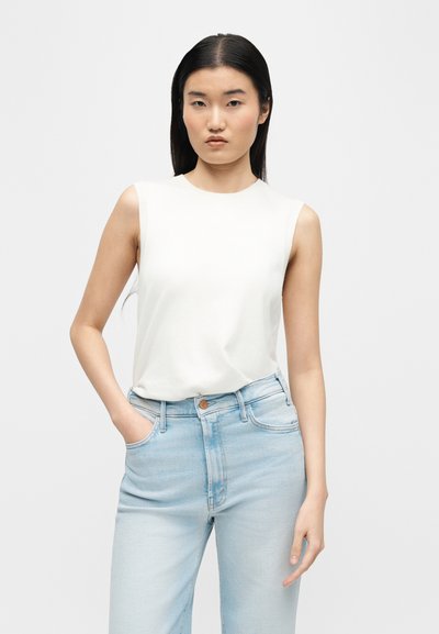 Γυναίκα με ίσια μαύρα μαλλιά φοράει λευκό αμάνικο τοπ και ανοιχτό μπλε ψηλόμεσο τζιν, στέκεται με το ένα χέρι στην τσέπη μπροστά σε λευκό φόντο.
