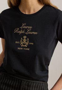 Μαύρο t-shirt με χρυσή επιγραφή "Lauren Ralph Lauren", έμβλημα με θυρεό, "EST. 1996" και "NEW YORK" κεντημένα μπροστά, φορεμένο με παντελόνι με ρίγες.
