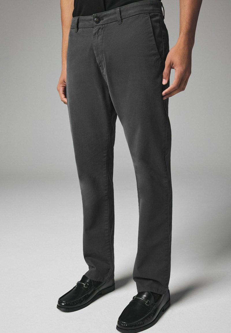 Graue Baumwollhosen mit schlankem Schnitt, ausgestattet mit einem Knopfverschluss, Seitentaschen und einer glatten Textur. Kombiniert mit schwarzen Slippern.