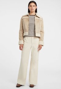 Giacca in shearling beige con design a colletto, abbinata a una maglietta a righe e pantaloni larghi color crema, caratterizzati da una superficie liscia e una vestibilità su misura.