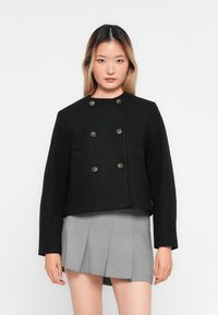 Jeune femme avec de longs cheveux portant un blazer noir à double boutonnage et une jupe mini plissée grise, se tenant devant un fond blanc uni.