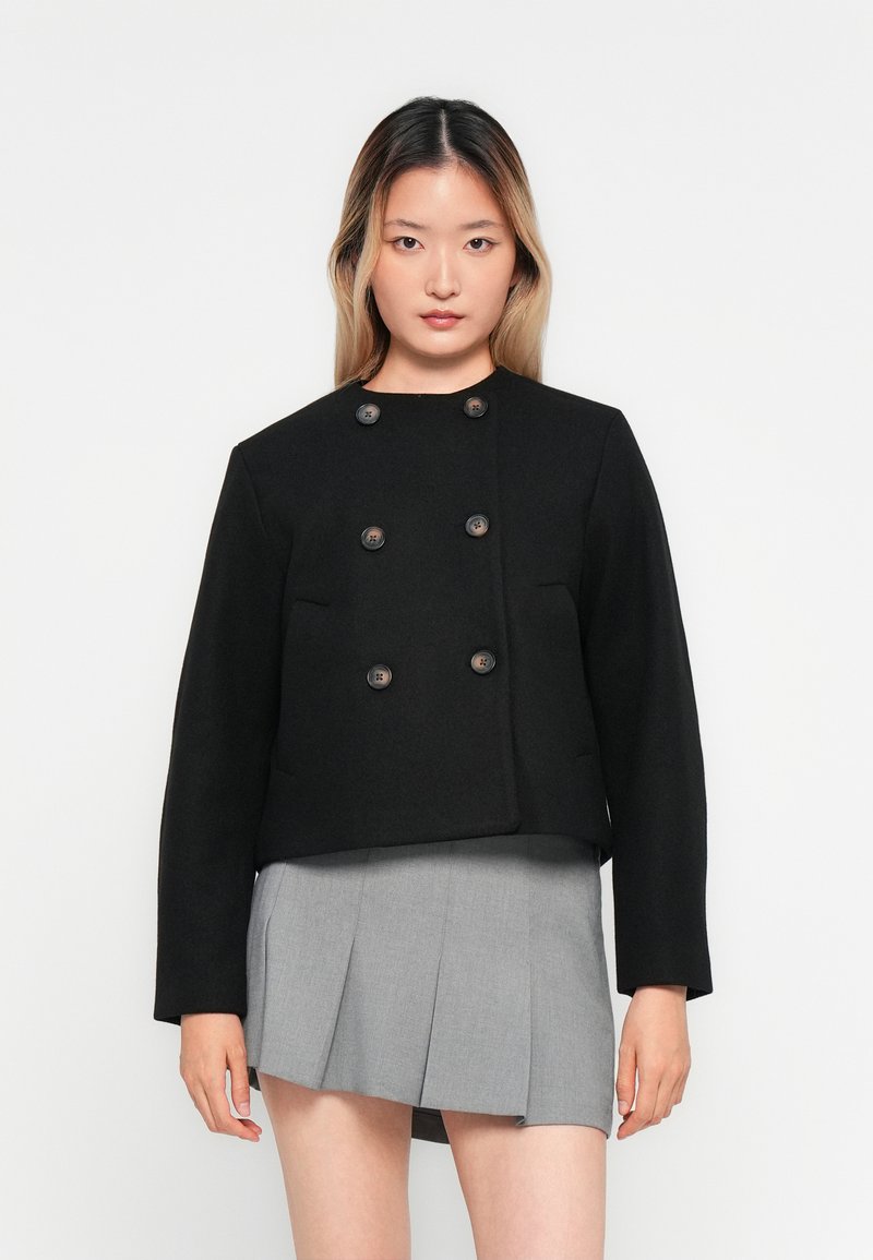 Jeune femme avec de longs cheveux portant un blazer noir à double boutonnage et une jupe mini plissée grise, se tenant devant un fond blanc uni.