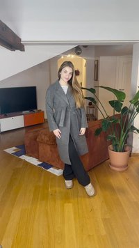 Casaco cinza de mistura de lã com cinto, design ajustado, mangas compridas e lapela com entalhe. Em pé sobre chão de madeira junto a uma planta e um sofá.