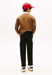 Bruine sweatshirt, zwarte cargo broek met zijzakken, witte sneakers en een rode pet. De outfit is casual met een relaxte pasvorm.