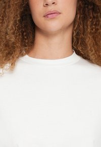 T-shirt blanc à col rond en tissu lisse. Présente un logo discret sur la poitrine et des manches courtes, avec une coupe décontractée.